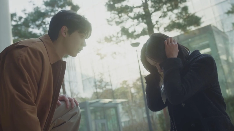 屋外で、男性が頭を抱えて座り込んでいる女性に寄り添い、心配そうに見守っている。感情的な瞬間を捉えたシーン。