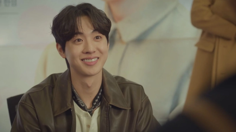 茶色のレザージャケットを着用し、明るい笑顔でこちらを見つめる若い男性のポートレート。背景はぼかされており、屋内での撮影と見られます。