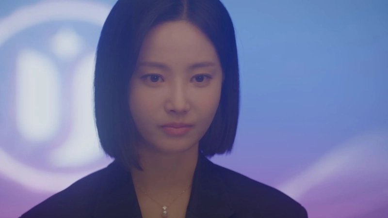 短い黒髪の女性が正面を見つめているポートレート。黒いジャケットを着用し、パールのネックレスを身につけています。背景は青と紫のグラデーションで、左側にぼんやりとしたロゴのような光が見えます。