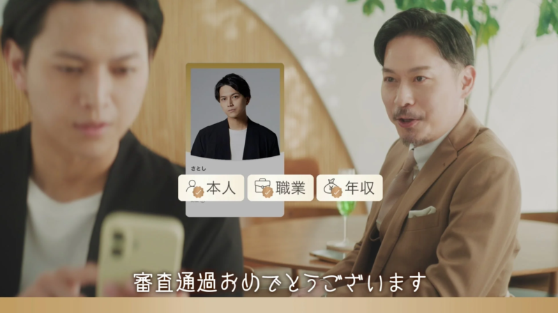 携帯電話を操作する男性と話している男性が映っており、中央には「さとし」という名前と写真、そして「本人」「職業」「年収」という情報を示すアイコンが表示されています。画面下部には「審査通過おめでとうございます」というメッセージがオーバーレイされており、何らかの審査に合格したことを祝う場面を示しています。