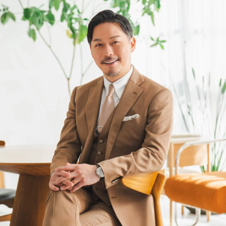 茶色のスリーピーススーツを着たアジア人男性が、明るい室内で椅子に座り、穏やかな笑顔でカメラを見つめています。