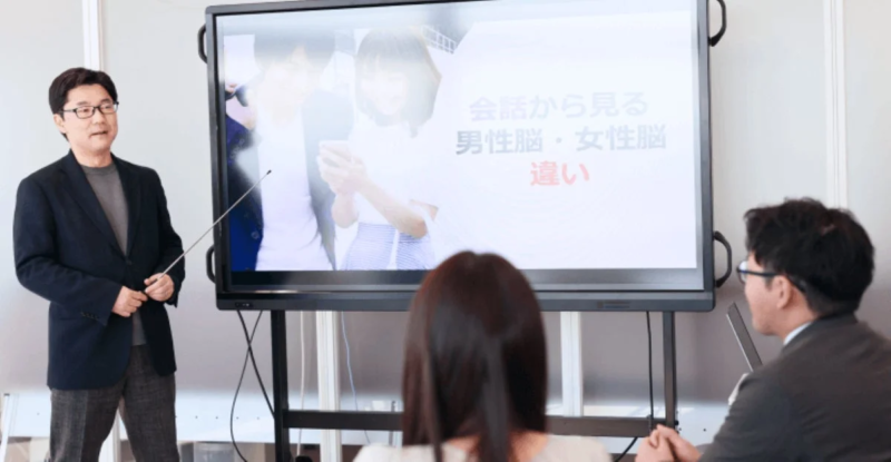 眼鏡をかけた男性が、大型ディスプレイを使って「会話から見る男性脳・女性脳の違い」というテーマでプレゼンテーションを行っています
