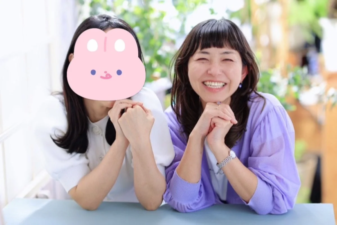 テーブルを挟んで座る2人の女性が写っています。一人は満面の笑みを浮かべており、もう一人の顔はピンクのウサギのマスクで隠されています。