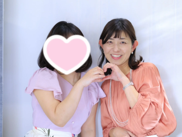 二人の女性が並んでおり、一人は笑顔で両手でハートのポーズを作っています。