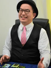 黒いハットと眼鏡をかけ、ピンクのネクタイとベストを着用した男性が、テーブルに座って笑顔を見せているポートレートです。手前には数枚のカードのようなものが置かれています。
