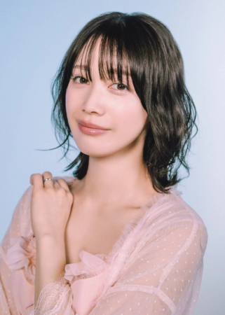 水色の背景を背に、ピンクのシアーなトップスを着たショートヘアの若い女性のポートレートです。彼女は穏やかな笑顔を浮かべ、左手には指輪をしています。