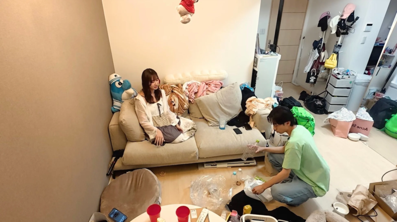 散らかったリビングルームで、女性がソファに座り、男性が床で片付けをしている様子