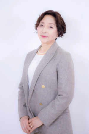 グレーのジャケットとパールネックレスを身につけた笑顔の中年女性のポートレート写真です。