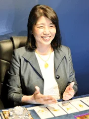 笑顔で話している東洋系の女性が写っています。彼女はビジネスウェアを着用し、テーブルに並べられたカードのようなものの前で、手でジェスチャーを交えながら何かを説明しているようです。
