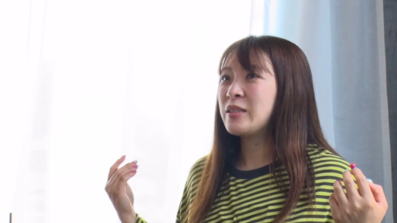 明るい背景の屋内で、ストライプ柄のトップスを着た若い女性が、何かを説明するように手を動かしながら話している様子の画像