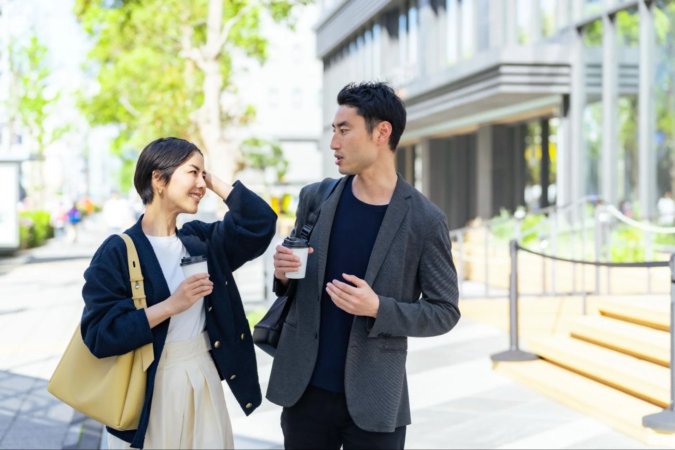 晴れた日の街中で、男女二人がコーヒーカップを片手に談笑しながら歩いています。