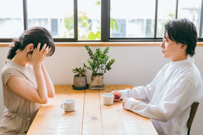 夫婦がテーブルを挟んで座っており、女性は顔を覆って悲しげな様子。男性は女性から視線を外し、二人の間に緊張感が漂っている。深刻な話し合いや関係性の問題を暗示する場面。